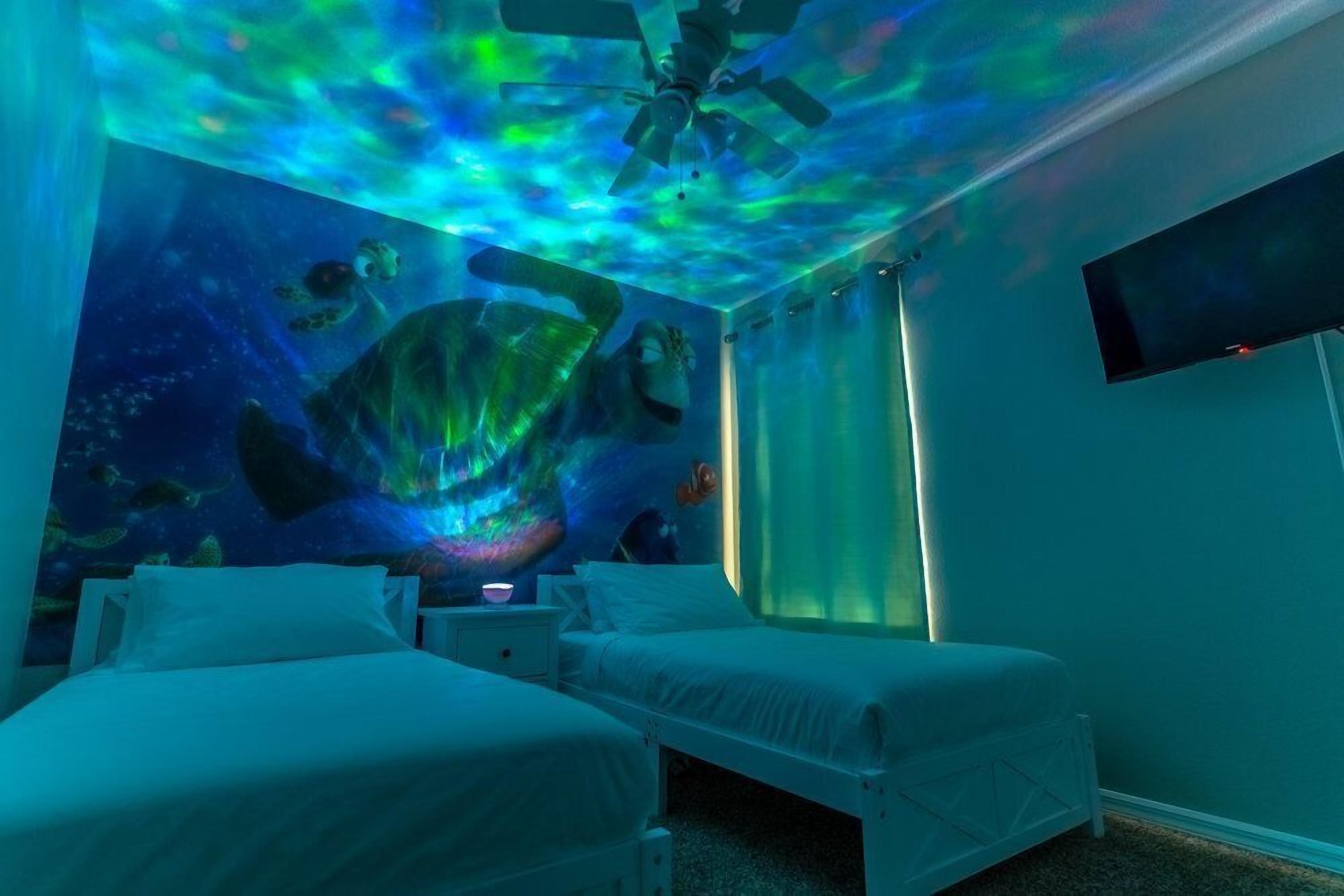 A room with two beds, a ceiling fan, a TV, and a mural of a turtle swimming underwater.
