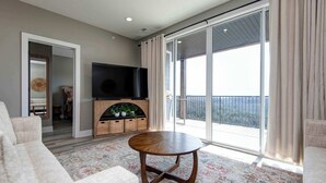 A modern living room with a round wooden coffee table, a flat-screen TV, and a large sliding glass door leading to a balcony. - BWV 24-6 Abiding Place (Branson)