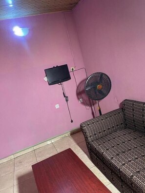Une pièce avec un mur rose, un canapé à motifs, une table en bois, un téléviseur fixé au mur et un ventilateur sur pied.