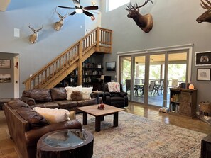 Ein geräumiges Wohnzimmer mit einer Ledersofa, einem Couchtisch und einem Bücherregal. Eine Holztreppe führt zu einem Balkon mit Sitzgelegenheit im Freien.