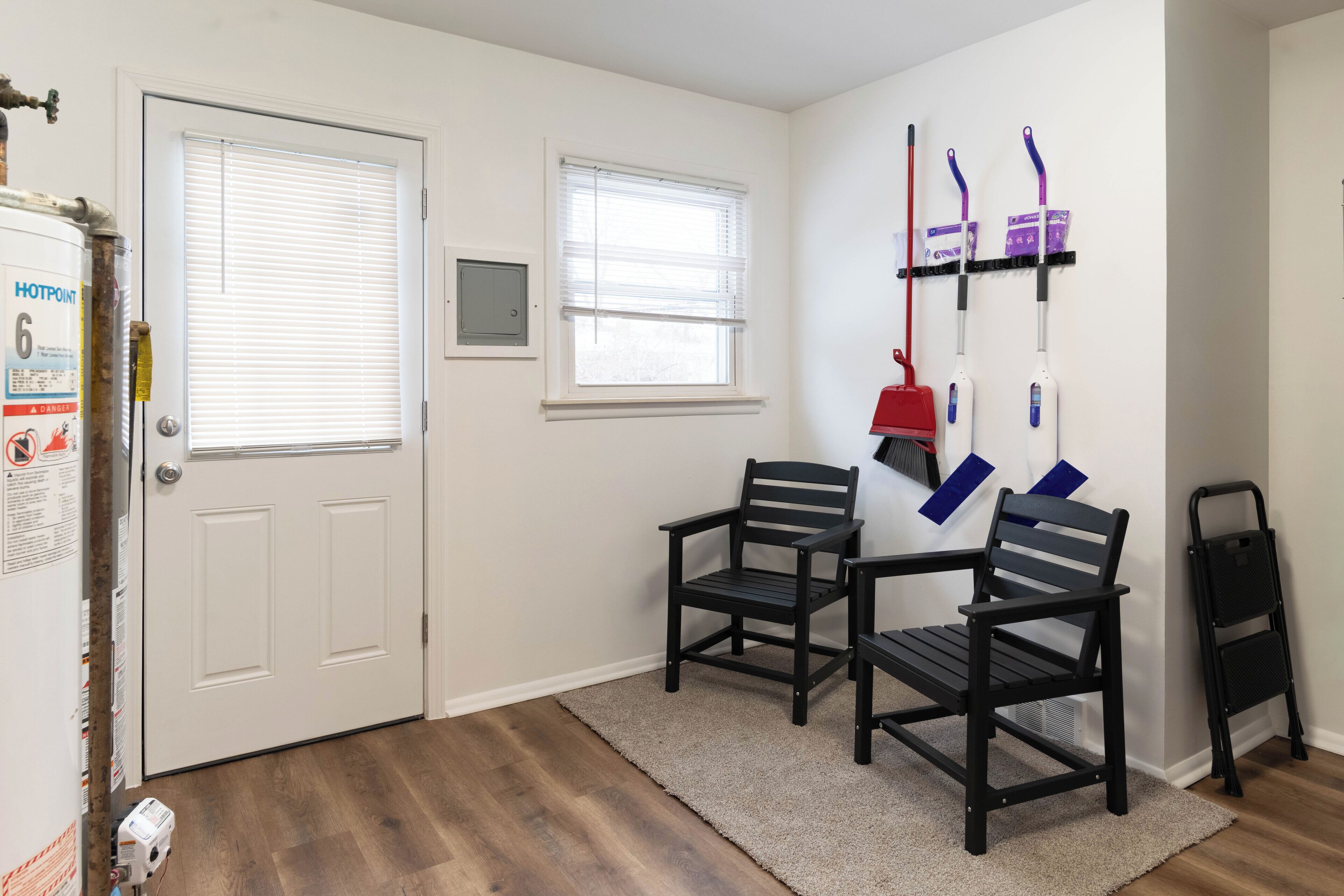 A room with a white door, two windows with blinds, a black chair, a red mop, a black ladder, and a fire extinguisher on the wall.