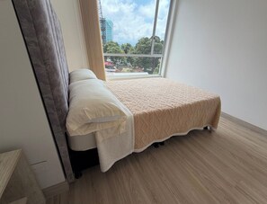 Un llit individual amb un edredó beix i almoades blanques en una habitació amb terres de fusta i un gran finestral.