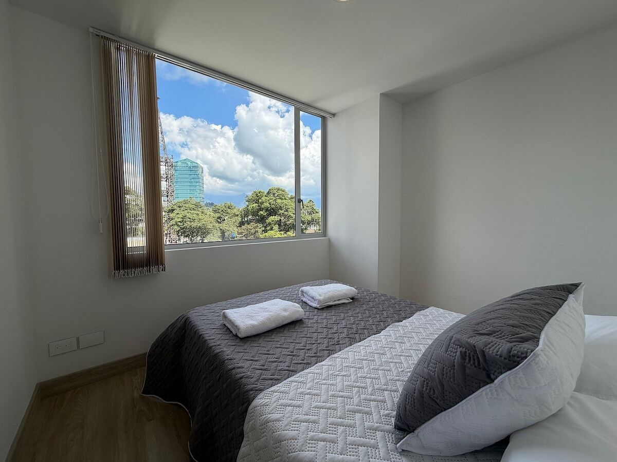 Un dormitorio con cama, ventana con cortinas y vistas a árboles y un edificio.