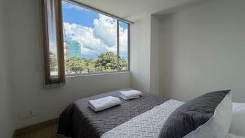 Un dormitorio con cama, ventana con cortinas y vistas a árboles y un edificio.