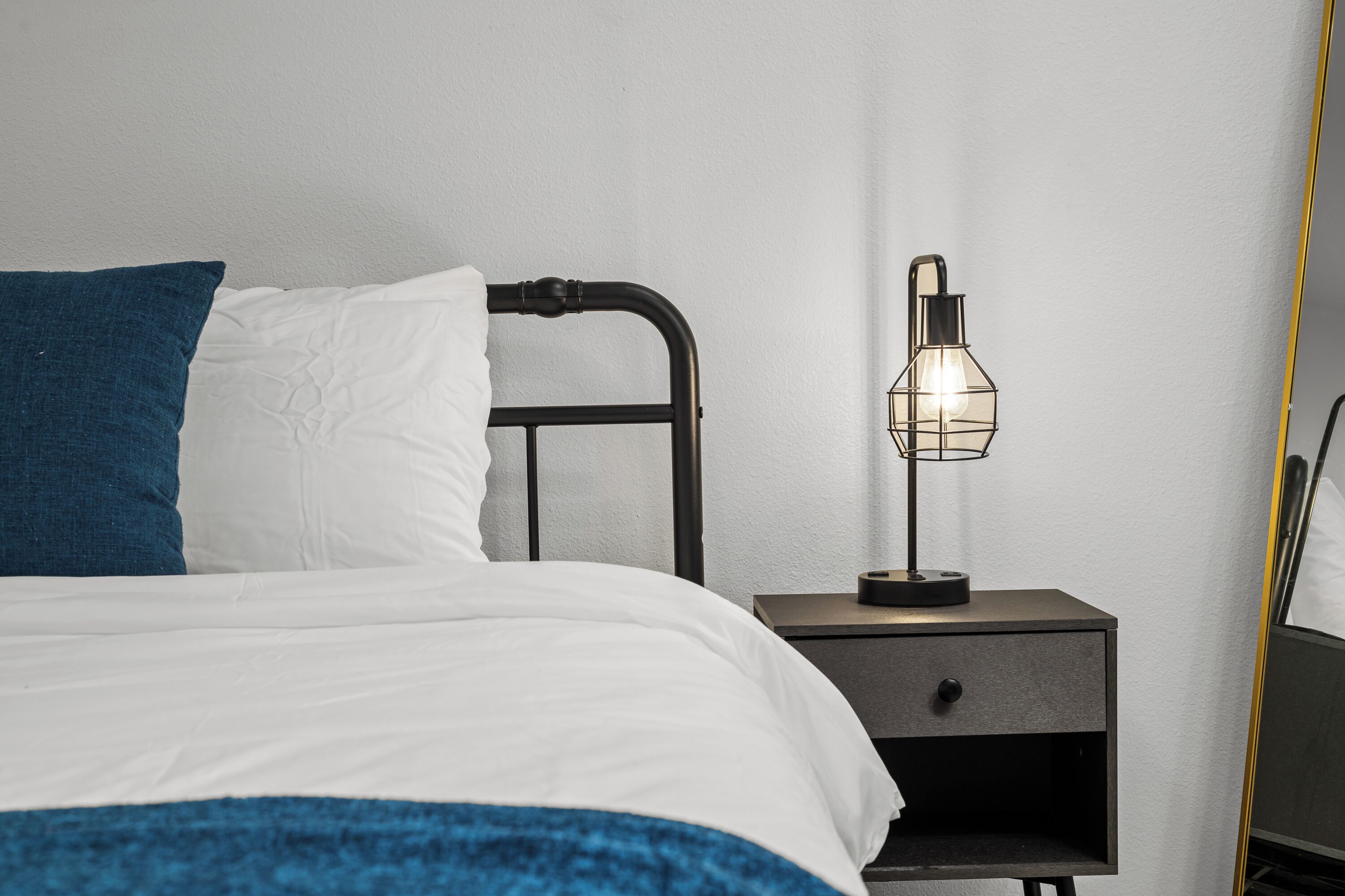 Una cama bien hecha con ropa de cama blanca y azul, un cabecero de metal negro y una mesita de noche con una lámpara.