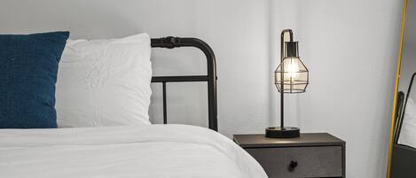 Una cama bien hecha con ropa de cama blanca y azul, un cabecero de metal negro y una mesita de noche con una lámpara.
