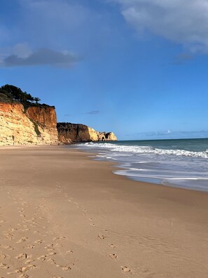 Uma praia com céu claro, ondas calmas e um penhasco rochoso com vegetação rasteira.