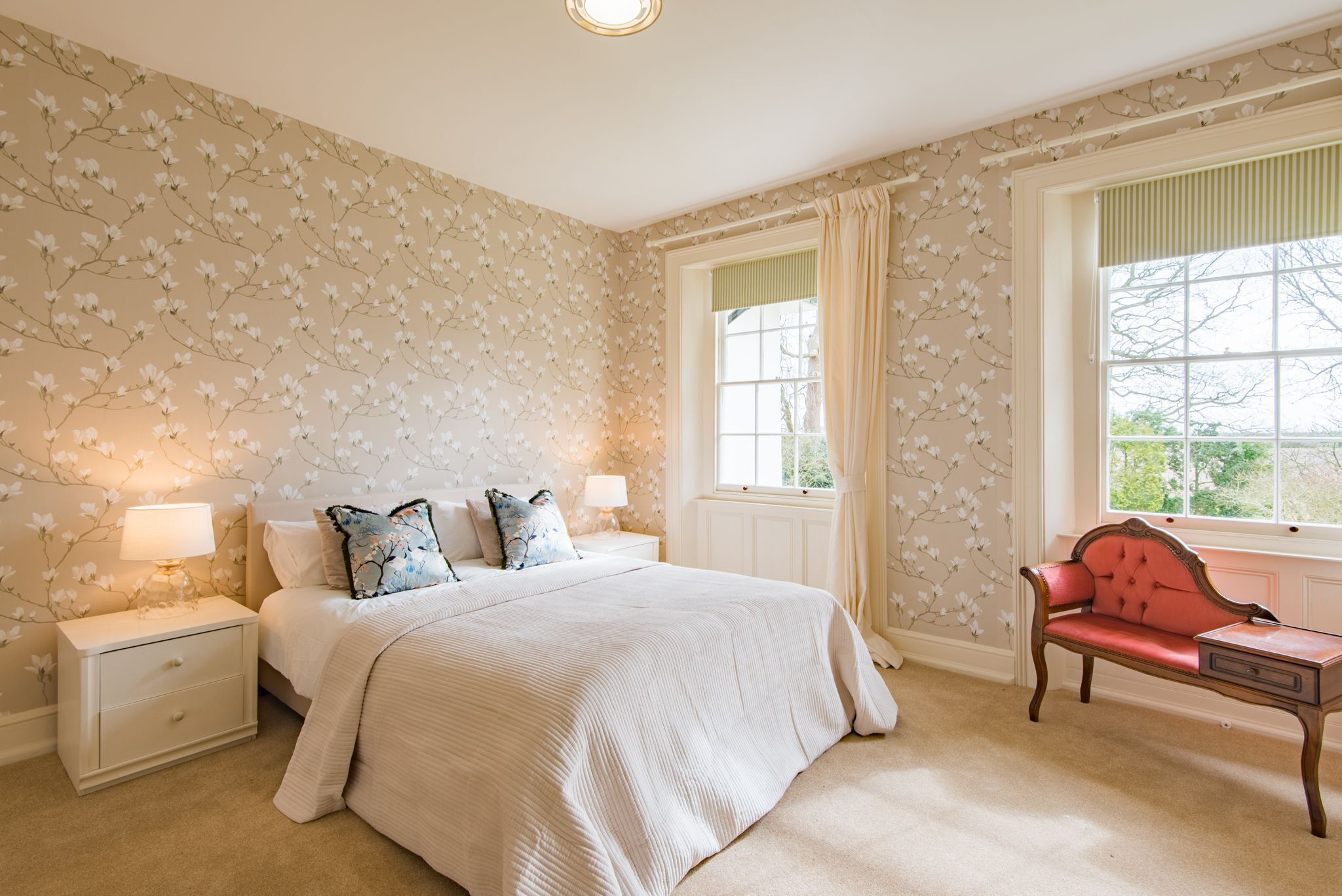 A bedroom with a large bed, two bedside tables with lamps, a window with curtains, and a red bench.
