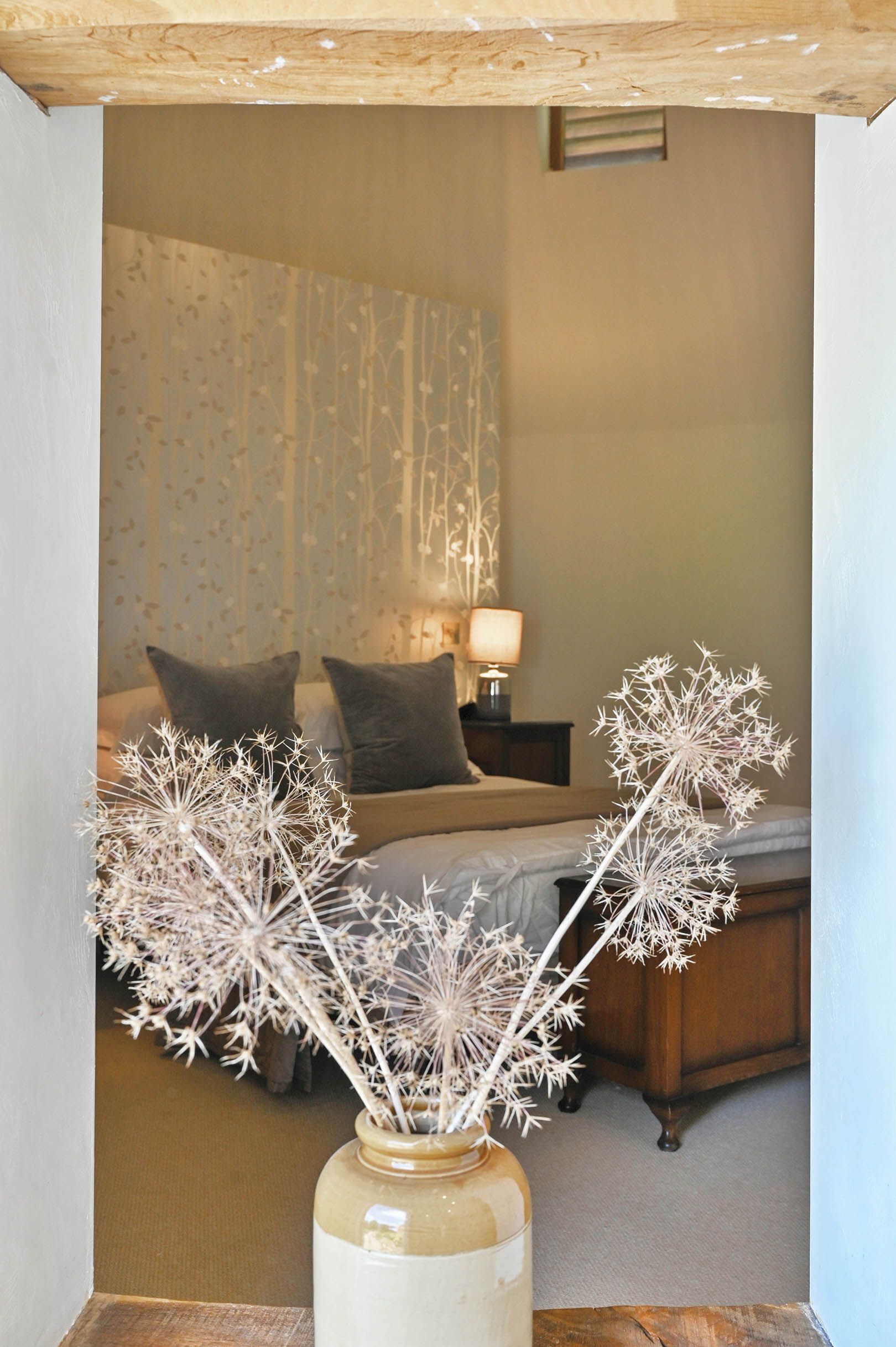 Ein Schlafzimmer mit einem Bett, Kissen, einer Nachttischlampe und einer Vase mit Trockenblumen.