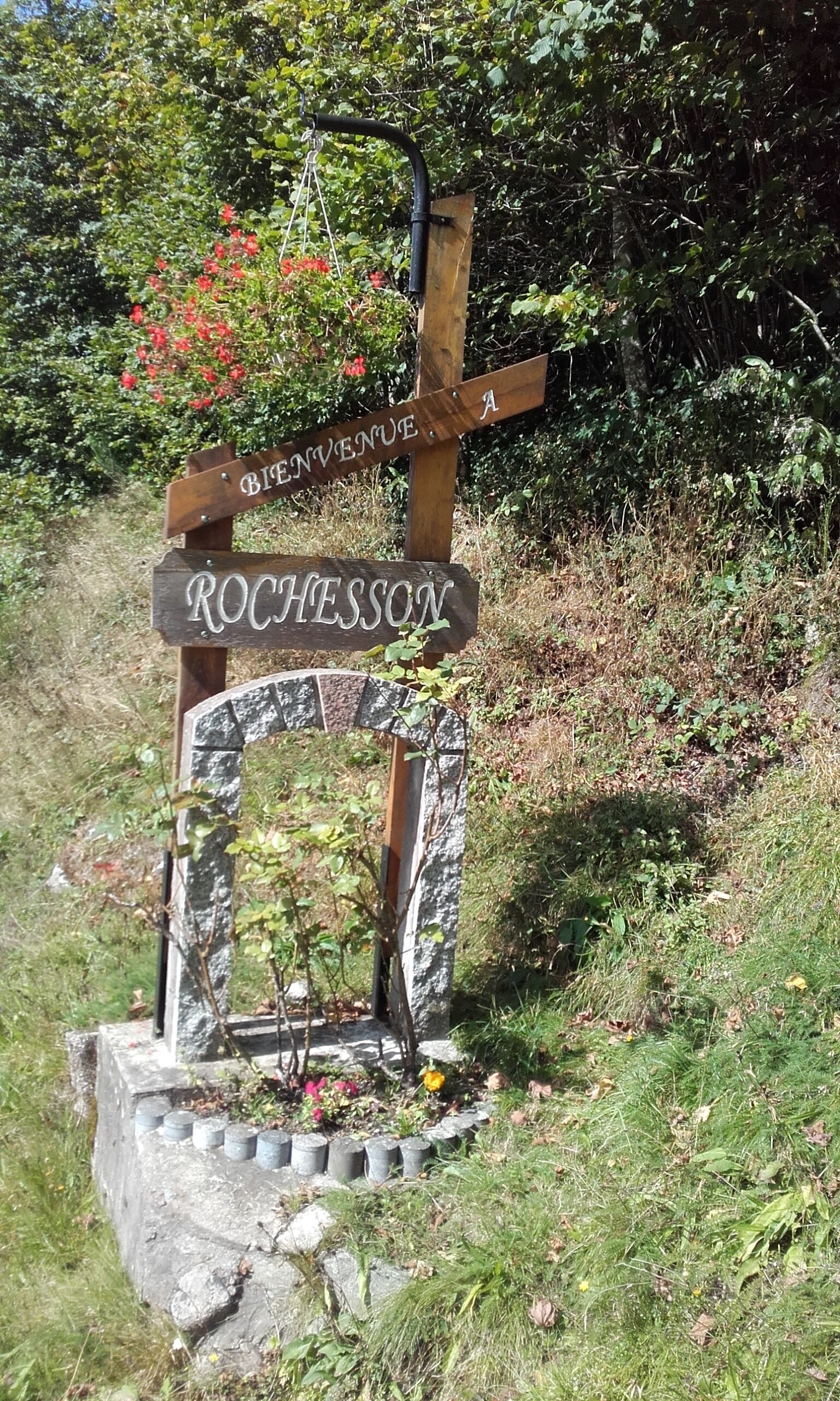 A wooden sign with 'ROCHESSON' and 'BIENVENUE A' inscribed on it, mounted on a stone base with a metal frame.