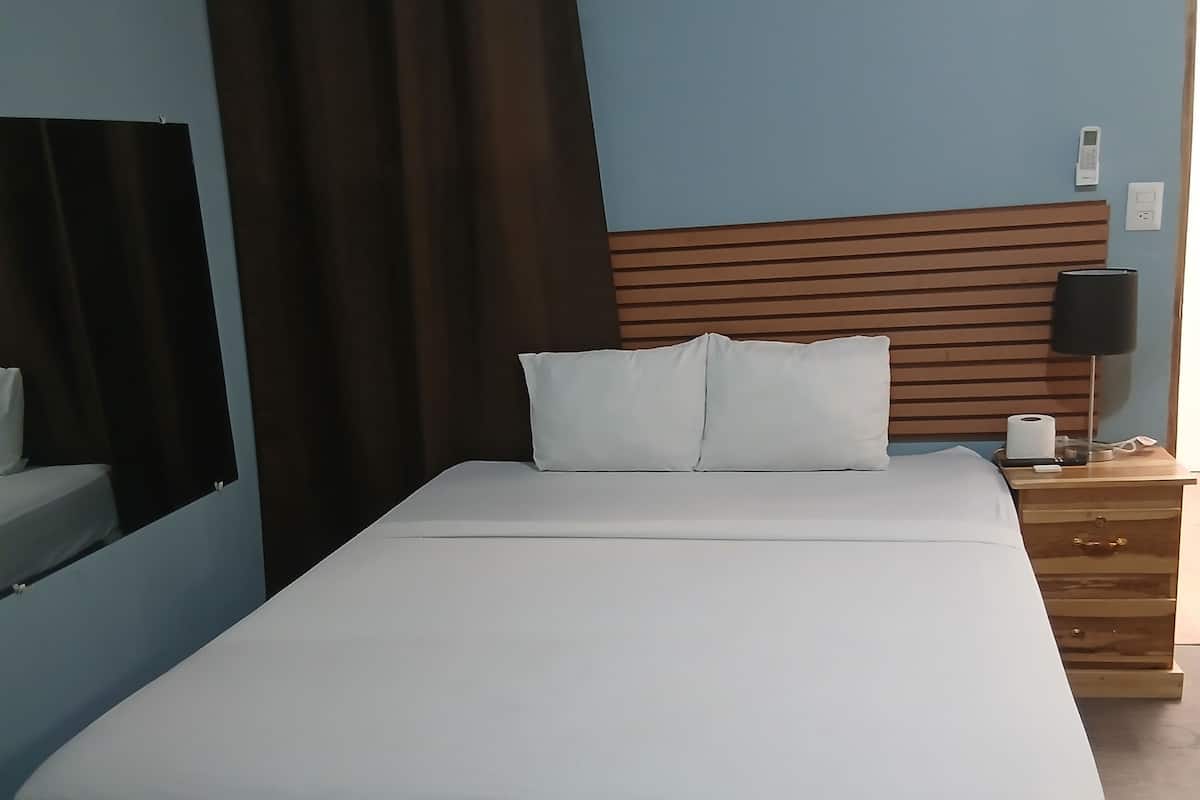 Una habitación de hotel estándar con una cama, una mesita de noche, una lámpara y un espejo.