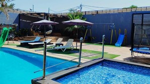 A swimming pool with lounge chairs and umbrellas, a slide, and a fenced area with palm trees. - Tala Villa - Dead Sea (Swemeh)
