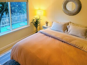 Ein Schlafzimmer mit einem Bett, einem Nachttisch mit Lampe, einem Spiegel und einem Fenster mit Blick auf einen Garten.
