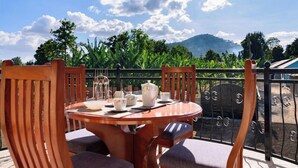 A round wooden table with chairs, a glass set, and a white pitcher on a balcony with a view of mountains and greenery. - Ngao Apartment & Suites Arusha (Arusha)