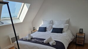 A neatly made bed with white and navy bedding, a bedside table with a lamp, and a window with a view of the sky. - Completely renovated Breton house - Walking distance to the beach - Sleeps 8 (Penmarch)