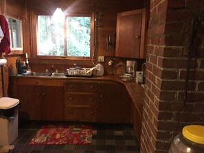 A kitchen with wooden cabinets, a sink, a window with a view of trees, and a brick wall. - Classic Waterfront Maine Cottage (Madison)
