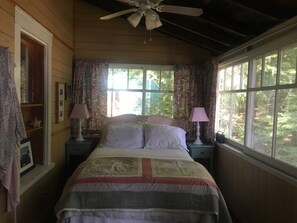 A bedroom with a bed, two bedside tables, a ceiling fan, and large windows with curtains. - Classic Waterfront Maine Cottage (Madison)