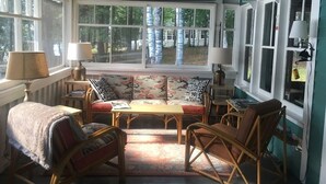 A cozy room with a wooden ceiling, a large window with white frames, a sofa with patterned cushions, and a small table with a lamp.
