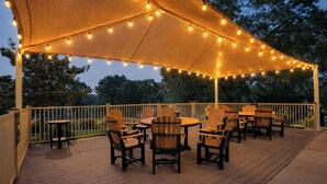 An outdoor patio with wooden chairs and tables under a canopy, string lights, and a white fence. - Modern Spacious Condo with no step access, pools, fishing, games and golf! (Branson)