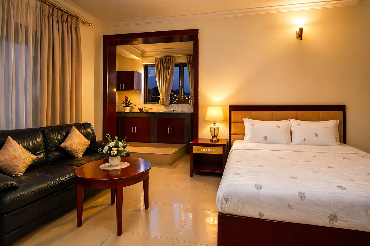 A hotel room with a bed, a sofa, a nightstand, a lamp, and a view of the city through a window.