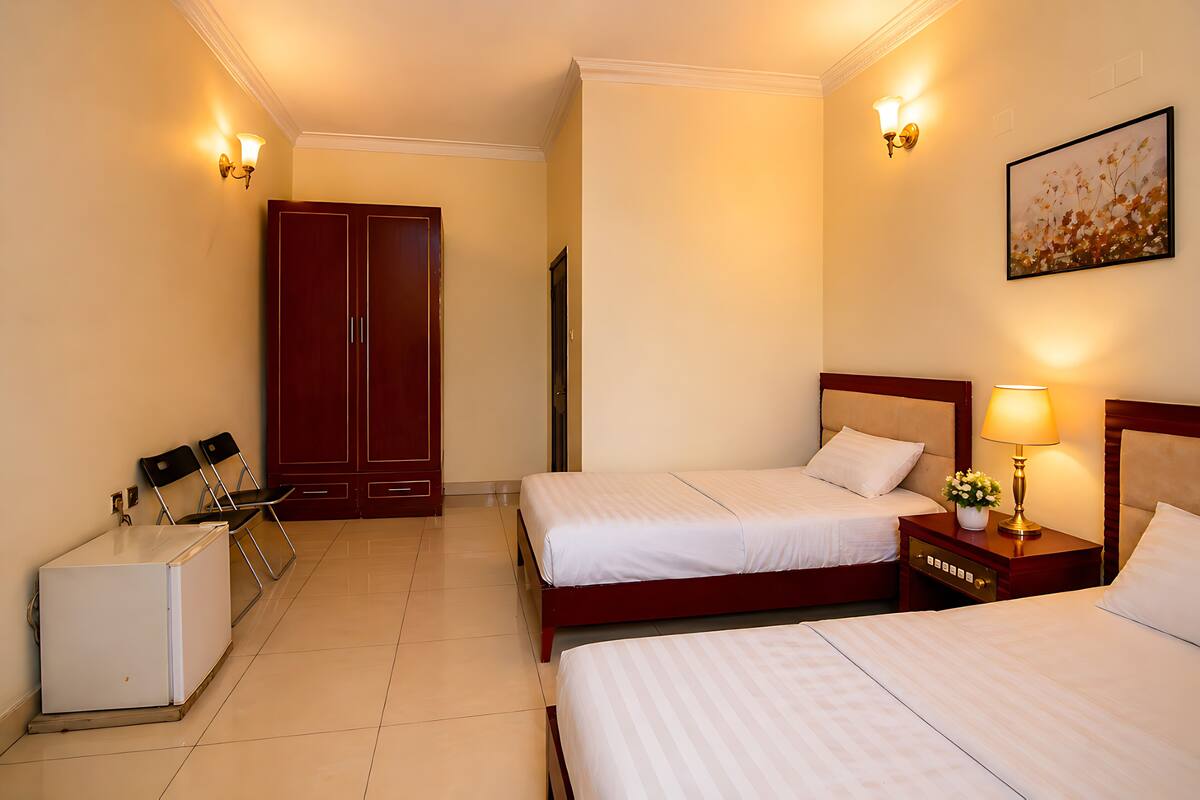 A hotel room with two beds, a wardrobe, a desk with a lamp, and a refrigerator.