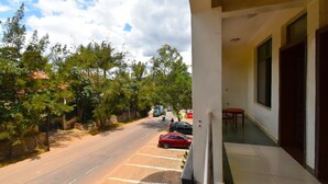 A view from a balcony overlooking a street with parked cars, trees, and a building with large windows and a door. - DV Appart Hotel (Kigali)