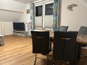 A dining area with a table, chairs, a TV, and a window with curtains. - Island star (Borkum)