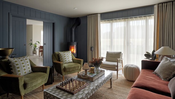 A living room with a fireplace, two armchairs, a sofa, a coffee table with a chessboard, and a large window with curtains.