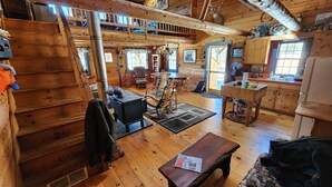 L’intérieur d’un chalet rustique est doté d’un escalier en bois, d’un espace de détente central avec une table et des chaises, d’un coin cuisine avec un réfrigérateur et d’une fenêtre qui laisse entrer la lumière naturelle.