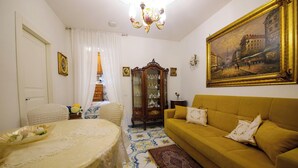 Una habitació amb taula de dinar coberta per un mantell de puntes, un sofà amb coixins, un penjador de cristall, un quadre a la paret i un paviment amb dibuixos.