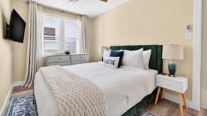 Et soveværelse med en seng, et natbord, en lampe, en loftventilator, et fjernsyn og et vindue med gardiner.