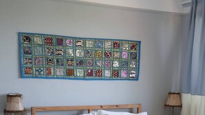 Un letto rifatto con lenzuola bianche, una testiera in legno e una coperta bianca. Al muro sopra il letto si trova una tappezzeria colorata, illuminata da un lampadario a sospensione.