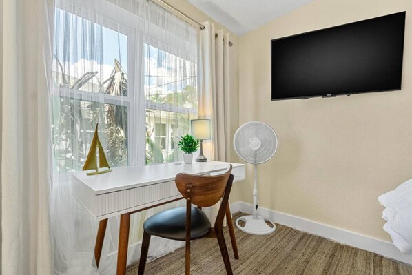 Une chambre d’hôtel avec un bureau blanc, une chaise, un ventilateur sur pied, un téléviseur fixé au mur et une fenêtre ornée de rideaux légers.