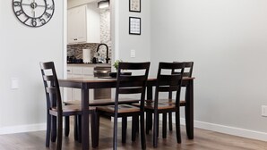Un coin repas avec une table et des chaises en bois, une horloge murale et une cuisine visible à travers une porte ouverte.
