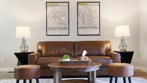 Un salon avec un canapé en cuir marron, deux petites tables basses rondes en bois et deux poufs assortis. Deux cartes encadrées sont accrochées au mur, et deux lampes sont posées sur des tables d’appoint.