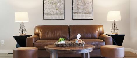 A living room with a brown leather sofa, two round wooden coffee tables, and two matching ottomans. There are two framed maps on the wall, and two lamps on side tables.