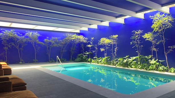 Ein moderner Poolbereich im Innenbereich mit klarem Wasser, blauen Wänden und einer Sitzecke mit braunen Kissen.