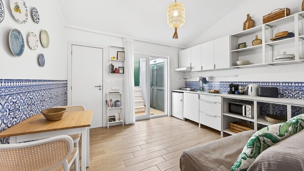 Eine moderne Küche mit weißen Schränken, ein Essbereich mit Holztisch und Korbstühlen sowie ein gemütlicher Wohnbereich mit einer gemusterten Decke auf der Couch.