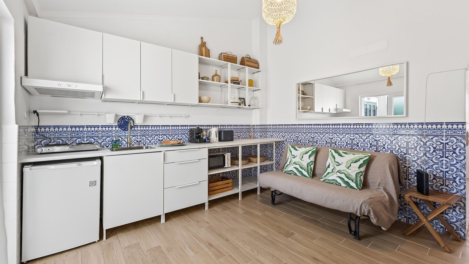 Una cocina funcional con armarios blancos, un refrigerador pequeño y un microondas. Hay un sofá con cojines estampados y una lámpara colgante.