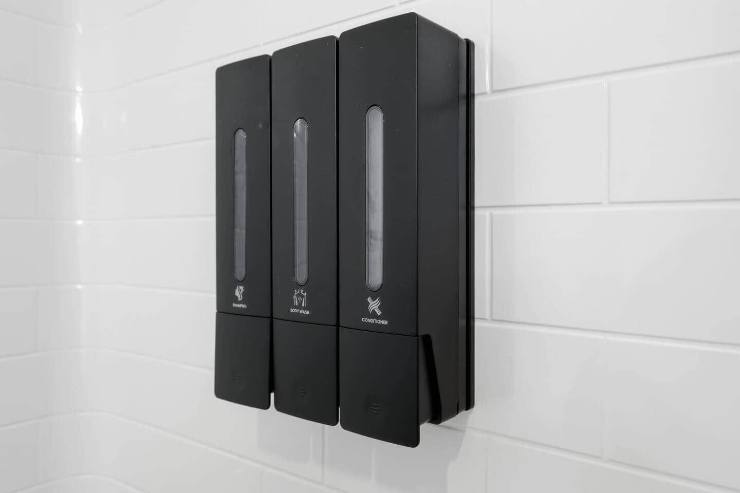 Een zwart, aan de muur bevestigd doseringssysteem met drie vakken, gelabeld voor shampoo, douchegel en conditioner.
