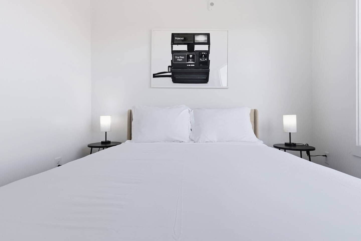 Habitación de hotel minimalista con una cama grande, dos mesitas de noche con lámparas y un cuadro enmarcado en la pared.