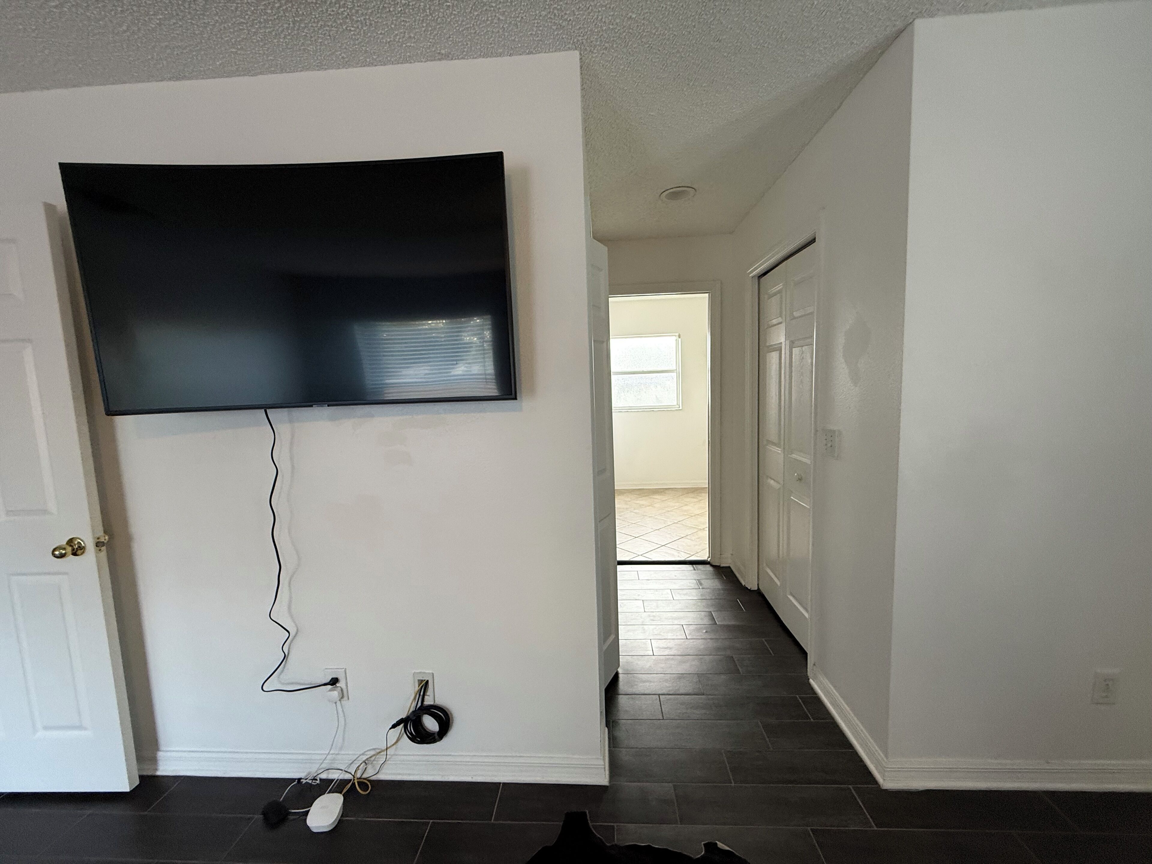 Ein Zimmer mit einem wandmontierten Flachbildfernseher, einer Tür, einem Flur und einem gefliesten Boden.