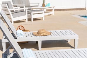 A poolside area with white lounge chairs, a table, and a straw hat.
