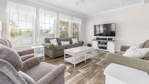 A living room with a sofa, coffee table, TV, and a ceiling fan. - Sunny Bungalow Retreat w/Pool + 6 Person Golf Cart Included (Emerald Isle)