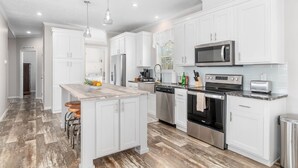 A modern kitchen with white cabinetry, stainless steel appliances, and a central island with stools. - Sunny Bungalow Retreat w/Pool + 6 Person Golf Cart Included (Emerald Isle)