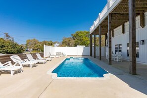 A swimming pool with lounge chairs, a covered patio, and a white building with windows.