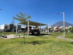 Un parque infantil con toboganes y una zona cubierta, rodeado de edificios modernos y una montaña al fondo.