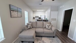 A living room with a sectional sofa, a patterned rug, a ceiling fan, and a kitchen in the background. - Cozy Beachy Riverwalk Condo – Close to Downtown & Beaches (Pensacola)