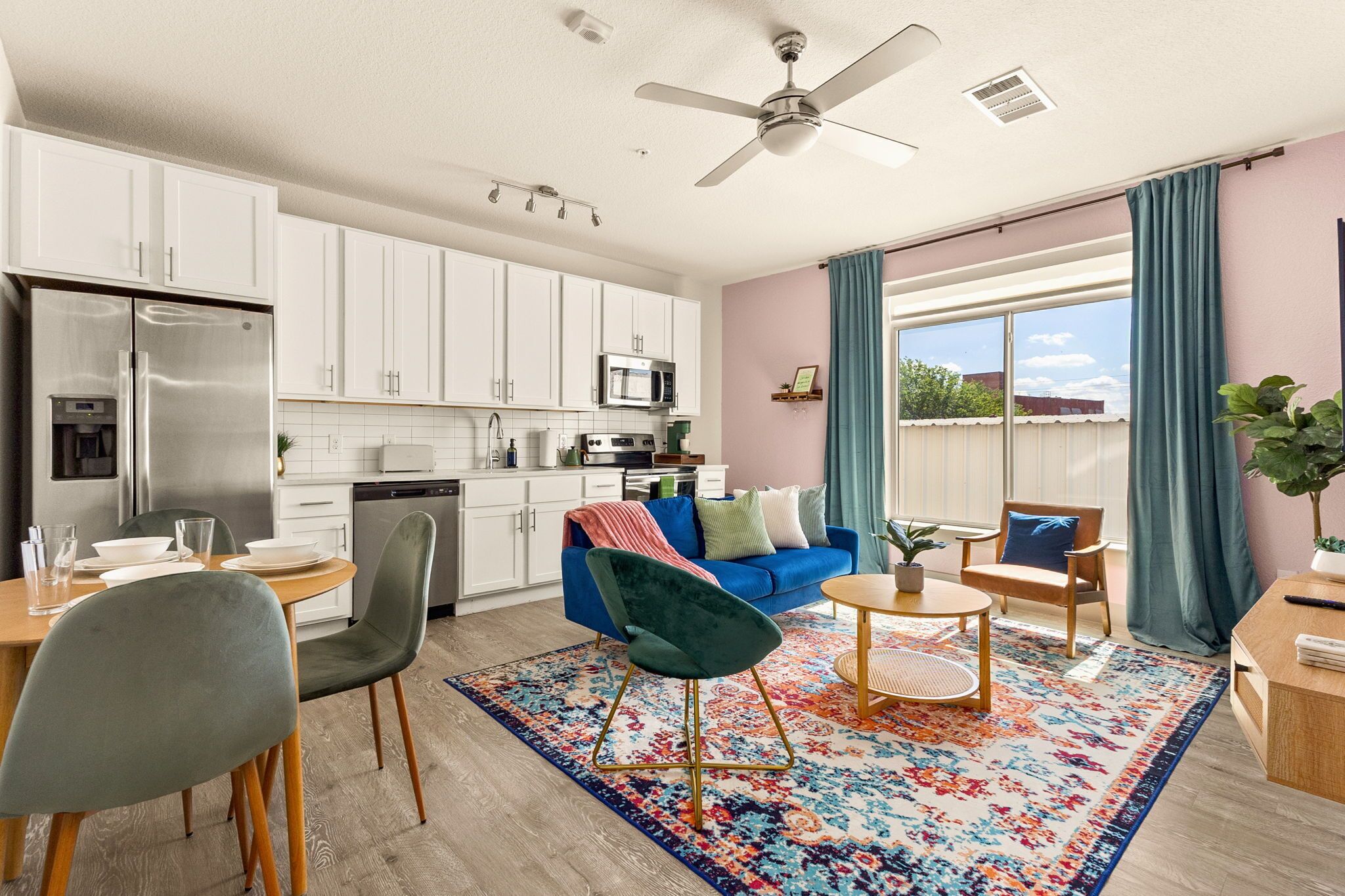 A modern kitchen with white cabinets, a stainless steel refrigerator, and a microwave. There is a dining area with a table, chairs, and a colorful rug. A blue sofa and a chair are in the living area.