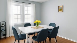 A dining room with a white table, four dark chairs, a vase of yellow flowers, a picture on the wall, and a black shelf with a plant.
